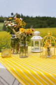 Bunte Wiesenblumensträusse in Vintage-Gläsern, Dekokäfig und Laterne auf gelbweiss gestreiftem Tischtuch in Freiem