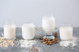 Various types of vegan milks in glasses with their ingredients (oat milk, coconut milk, almond milk and rice milk)