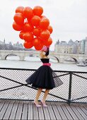 A young woman on a bridge wearing a black petticoat dress with a pink belt and holding a bunch of red balloons