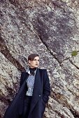 A short-haired woman standing in front of rocks wearing a dark virgin wool coat and a striped shirt