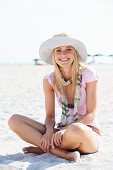 Junge blonde Frau mit Sommerhut, Schal und rosa T-Shirt sitzt im Sand am Strand