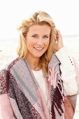 A blonde woman on a beach wearing a pink-toned shawl over her shoulders