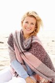 A blonde woman on a beach wearing a blue jumper and a pink-toned shawl over her shoulders