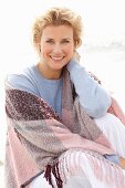 A blonde woman on a beach wearing a blue jumper and a pink-toned shawl over her shoulders