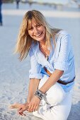 A young blonde woman on a beach wearing a light-blue shirt and white trousers