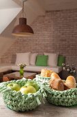 Fruit and bread in green crocheted baskets on wooden table in front of seating area against brick wall