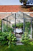 Seating area with white tablecloth on Tulip Table and garden plants in greenhouse with large foliage plants around door