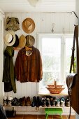 Jackets and hats hung from coat pegs on white wooden wall above full shoe rack below window