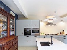 Antique, glass-fronted cabinet in open-plan, white kitchen with designer pendant lamp