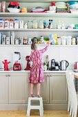 Girl on step stool in front of kitchen counter and open shelf full of storage containers
