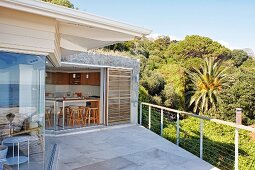 Spacious terrace adjoining house with view into kitchen through open sliding door; tropical forest in background