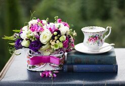 Romantisches Blumengesteck mit pinkfarbenem Schleifenband neben antiquarischen Büchern und nostalgischer Tasse auf Vintage Koffer