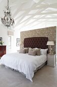 Pale bedroom area with wallpapered partition, dark brown, button-tufted headboard, chandelier and white-painted roof structure
