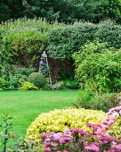 Flowering shrubs, clematis climbing through obelisk, lawn and wall in well-tended garden