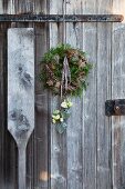 A wreath of pine cones, green twigs, moss and wooden stars with Japanese roses in a hanging vase against a weathered wooden door