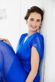 A brunette woman wearing a blue dress sitting against a wooden wall