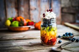 A colourful fruit salad in a jar topped with cream and pomegranate seeds on a rustic wooden surface