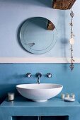 weiße Waschschüssel auf hellblauem Unterbau vor gleichfarbiger Wand, darüber runder Spiegel mit Vogelmotiv und romantischer Dekokette