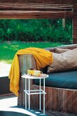 Wooden bench with seat cushions and yellow blanket in front of rolled up bamboo blind