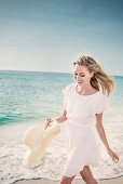 A blonde woman running along a beach wearing a white dress and holding a sunhat