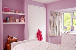 Child's bedroom with pink-painted walls, floating shelves in niche, white fitted wardrobe and chest of drawers below window