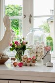 Romantic Easter arrangement on white windowsill; blown eggs in glass jar and bouquet of pink roses next to china rabbit