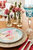 Romantisches Gedeck mit Vogelmotiv auf Teller und bunte Kristallgläser neben Glasfläschchen mit Rosen