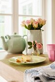Plate of cake, vase of roses and teapot on table