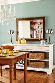 Antique mirror and table lamps on sideboard against pastel-blue wall: crystal chandelier and dining area in foreground