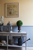 Lanterns and small bushes decorating console table against wall painted pastel yellow with pastel-blue wainscoting