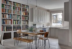 Square rustic wooden table, vintage chairs and bookcase in front of white fitted kitchen with panelled doors