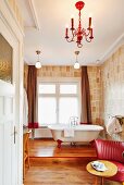 Free-standing bathtub on wooden platform behind table and red retro armchair in bathroom with designer wallpaper