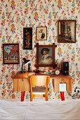 View across bed to 50s-style desk and chair below collection of pictures on floral wallpaper