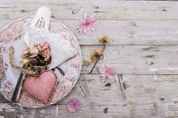 Pink heart-shaped ornament and posy of dried roses on vintage-style plate on rustic wooden surface scattered with flowers