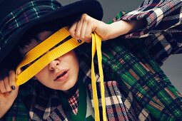 A young woman wearing an outfit made from various checked fabrics holding a yellow ribbon over her eyes