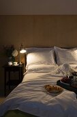 Brennende Nachttischlampe neben Doppelbett mit weisser Bettwäsche, im Vordergrund Frühstückstablett