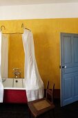 Vintage bathtub with shower curtain hanging from metal frame in rustic bathroom with yellow wall and blue door