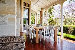 White wicker chairs at wooden table on reddish wooden floor of veranda with white wooden pillars