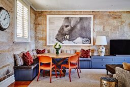 Comfortable corner of living room with orange plastic chairs around wooden table and blue fitted corner bench against stone wall