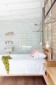 Double bed with wooden frame below pendant lamps and against brick wall in front of glass partition screening shower area with white-tiled wall