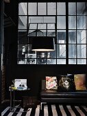 Side table next to black leather couch and arc lamp in front of Industrial interior window in masculine loft apartment