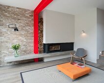 Low orange coffee table on rug and designer easy chair in front of grey fireplace; red steel structure and stacked firewood against brick wall