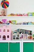Puppenhaus und rosafarbenes Puppenschloss auf weißem Regalschrank mit grünen Aufbewahrungsboxen, darüber Spielzeugsammlung auf weißen Wandboards
