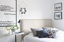 Scatter cushions on bed with fabric headboard, bedside lamp on bistro table and black and white framed motto on wall