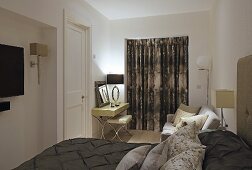 Elegantes Doppelbett mit dunkelbrauner Tagesdecke und drapierten Kissen, Schminktisch vor Fenstertür mit geschlossenem Vorhang