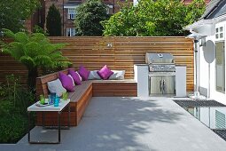 Eingebaute Holzbank mit violetten Kissen und Grill auf sommerlicher Terrasse, moderner Sichtschutzzaun