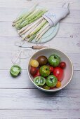 White and green asparagus on a wooden board, asparagus peelings, a peeler and a bowl of various tomatoes