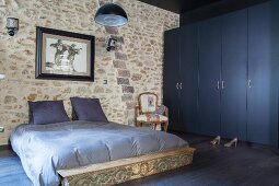 Grey-blue bed linen on double bed against stone wall next to dark blue wardrobes