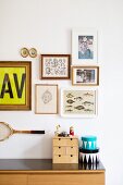 Miniature chest of drawers and boxes on top of sideboard below gallery of pictures and tennis racquet on wall