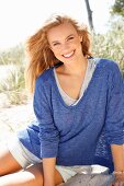 A young woman wearing shorts and a blue mottled shirt sitting on a beach